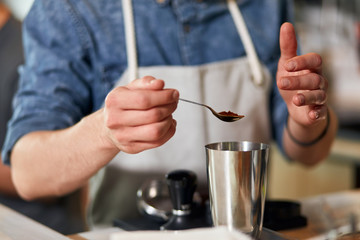 Fototapeta na wymiar Hands of male beared barista, keep little spoon with ground coffee over metal pot, measure coffee for cup of fresh aromatic cappuccino in coffee house