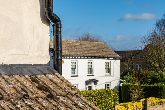 Shallow focus of part of a cottage roof and guttering in the foreground. The distant shows a nearby white painted cottage facia and hedged front during spring.