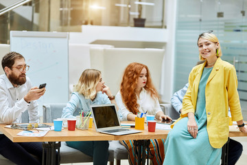 Beautiful young woman wears bright colourful clothes, sits on table with charming smile, looks at camera with positive expression, on background of busy colleagues