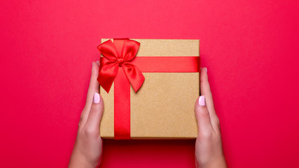 Woman manicured hands holding red and golden wrapped present or giftbox on deep red background, copy space, top view, flat lay. Background for Valentine's Day, Mother's Day.