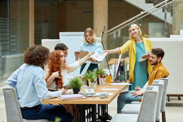 Caucasian puzzled young girl, has foxy hair and red lips, sits at table with designers with open mouth, stretches hand to take paper documents from peaceful leader, looks up at ceiling right side shot