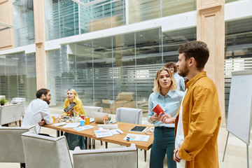 Pretty charming girl with long blonde hair, wears jeans and light blue t shirt, talks to handsome guy in casual outfit, looks away with open mouth, tells business cases in fashionable agency
