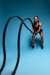 ambitious girl making a wave with battle ropes, full length photo. isolated blue background, studio shot. girl leads healthy active lifestyle