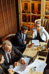 Down shot of business meeting, smart colleagues discussing business on the agenda at long table full of papers, documents and electronic devices in modern small office