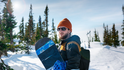 young charismatic bearded snowboarder portrait in snowy forest in winter sunny day. Man holding his snowboard and posing on top of the hill.