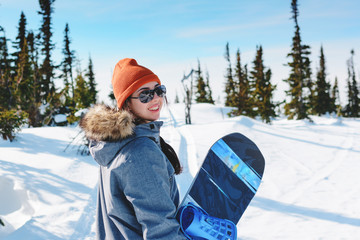 young charismatic girl snowboarder portrait in snowy forest in winter sunny day. Woman holding her snowboard and posing on top of the hill.