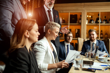 Close up of young smiling businesswoman reading paper documents at small crowded office on the background of standing men