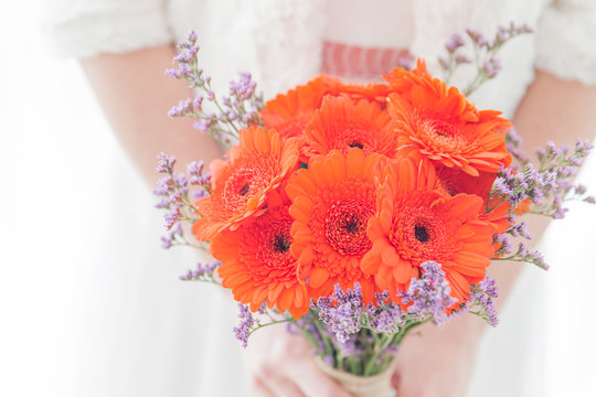 Close-up of a bride in a white lace dress holding a vibrant bouquet of orange gerbera daisies and purple statice flowers