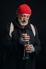 Happy old beardy man wearing red hat and black coat looking with hope at bottle of drinking. Isolated over black background