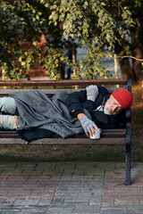 Vagrant male lying on street bench asking for money, for any help. Desperate and lonely homeless man without shelter