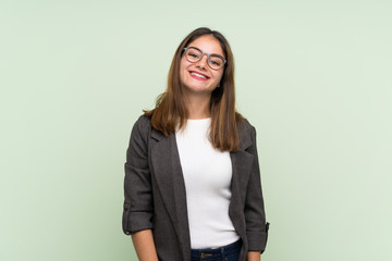 Young brunette girl with blazer over isolated green background with glasses and happy