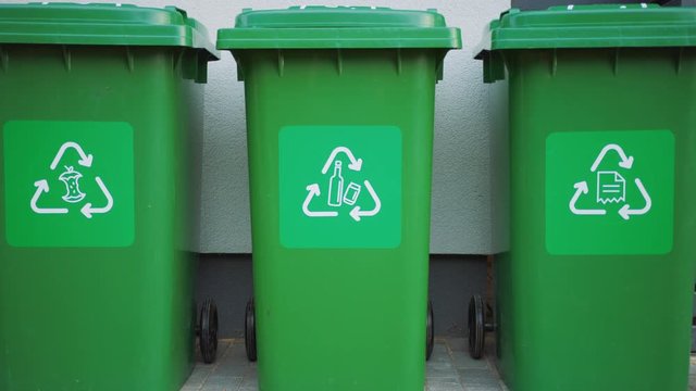 Green colored, plastic garbage bins, with different recycle logos on front, stacked in row against brown wood wall. Concept of waste sorting for food, paper and bottles. Saving Environment from Trash.