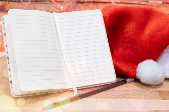 notebook,red Christmas hat and pencil on wooden table on background christmas garland