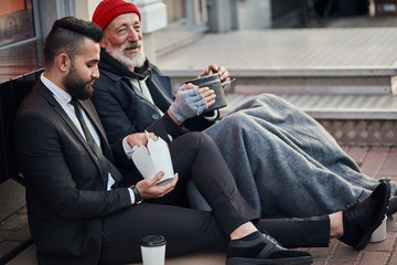 Handsome businessman in suit sitting on floor with homeless man together, listen to his story of...