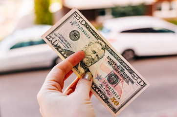 Female hand holds 100 dollar bill against the background of the street. US 50 dollar bill close up,...