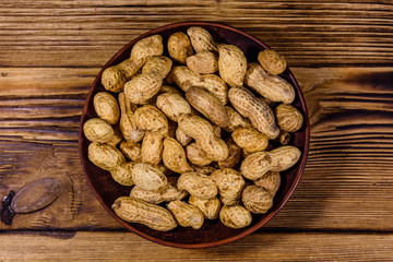 Ceramic plate with heap of unpeeled peanuts on wooden table. Top view