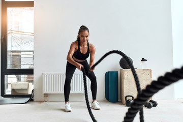 Concentrated Caucasian female doing crossfit exercises using rope in light gym. Young beautiful...
