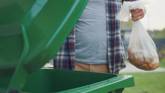 Close Up Shot of a Caucasian Male in Checkered Shirt Throwing Away Biological Food Waste into a Green Trash Bin. He Uses Correct Garbage Bin Because He is Sorting Waste and Helping the Environment.