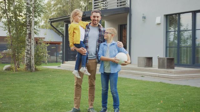 Portrait of a Happy Family of Three: Father, Daughter, Son. They Are Posing In Front of Camera on a Lawn Next to Their Country House. Dad is Holding the Girl in His Arms. Boy is Holding a Football.