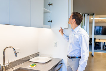Modern office kitchen room in building with man opening shelf cabinet by faucet looking inside