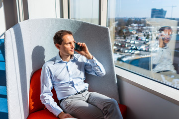 New modern office building with man talking on phone on chair by glass window looking through it happy businessman sitting with corporate view