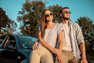 Beautiful travelling couple resting on car hood lit by sunset rays
