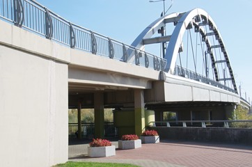 Fototapeta premium Against the blue sky new road bridge across the river Mukhavets. On the banks of the flower beds with red flowers. Near the Brest fortress. Built in Brest. Belarus.