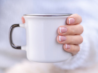 Woman in a cozy knitted sweater holds a winter cup close up. Female hands mockup with elegant french manicure nails holding a white ceramic mug with copy space. Winter and Christmas time concept