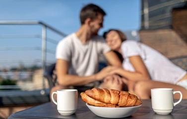 Morning coffee and croissants on a background of blurred young loving couple on balcony