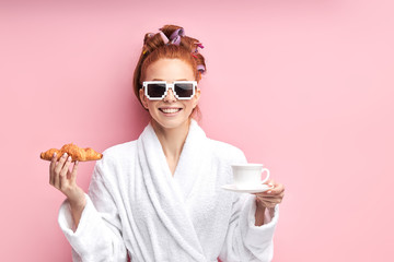 Caucasian girl after shower enjoying cup of tea with croissant. Isolated over pink background