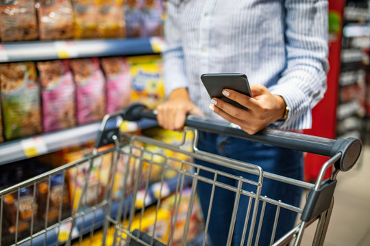 Shopping list. Woman with smartphone in store. grocery shopping. gadgets and shopping.  Picture of young cheerful woman in supermarket with shopping trolley choosing products and using phone.