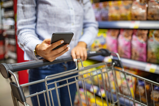 Woman using mobile phone while shopping in supermarket, trolley. Woman using mobile phone to compare price. Young woman shopping purchase healthy food in supermarket blur background.