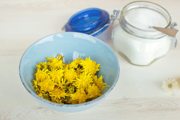 Washed yellow dandelion flower heads in blue bowl, jar with sugar and spoon on the wooden table. Ingredients for dandelion jelly. 