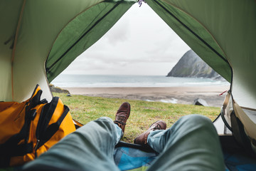 POV view from tent on on legs wearing hiking shoes in front of mountains and ocean. Alone tourist resting in camping by the sea - obrazy, fototapety, plakaty