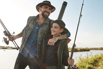 Father and son go to fishing place. They happy, smile. Father hugs son. They hold fishing rods. The sun shining.