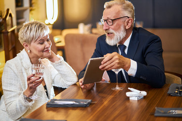 Attractive senior couple use modern technologies, tablet in restaurant. Grey-haired man in tuxedo, glasses and woman in white blazer