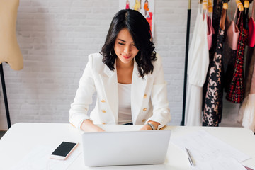 Contemporary confident Asian lady entrepreneur in stylish jacket working on laptop with garment...