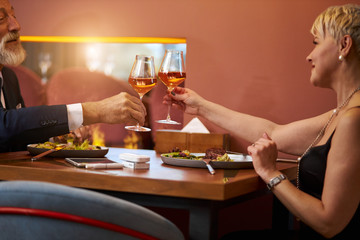 Grey-haired man in tuxedo and senior lady in black dress clink glasses with champagne in expensive restaurant. Look and happily smile. Romantic atmosphere
