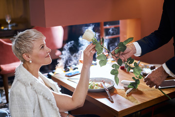 Pretty caucasian woman gets white rose as a present. Close up photo. Tablet, electronic cigarette, food on table. Pleasant moment. Love concept