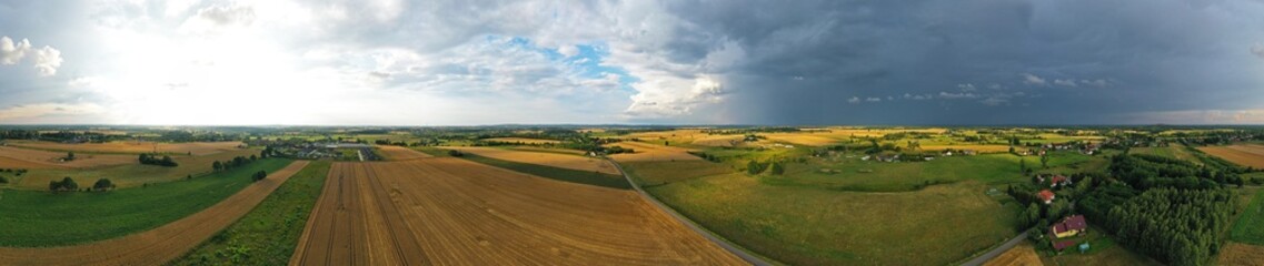 Aerial drone wide panoramic view on agricultural landscape with wheat field, village, meadows, forest and rain storm on horizon.