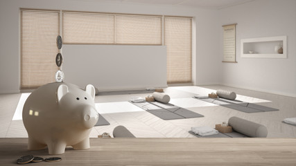 Wooden table top or shelf with white piggy bank with coins, empty yoga studio, open space with mats and accessories, ready for practice, renovation restructuring concept architecture