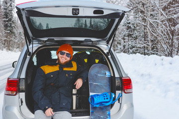 Happy snowboarder is sitting in back trunk of his car on the roadside in winter. Way to ski resort. Winter vacation concept. Healthy lifestyle.