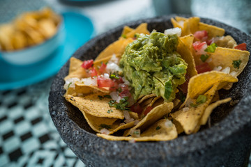 Yellow corn nacho chips garnished with ground beef, guacamole, melted cheese, peppers and cilantro leaves a molcajete, traditional Mexican mortar.