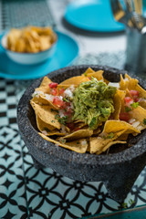 Yellow corn nacho chips garnished with ground beef, guacamole, melted cheese, peppers and cilantro leaves a molcajete, traditional Mexican mortar.