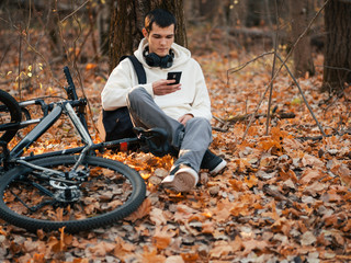 A young handsome male cyclist rests by a tree at sunset among the fallen autumn leaves in the Park.