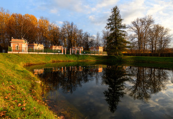 Fototapeta na wymiar Beautiful view of Kuskovo park in Moscow at sunny autumn evening with lush grass, yellow leaves and pond with reflection of blue cloudy sky, pine tree and bird houses. Kuskovo, Moscow