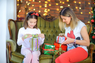 Mom and daughter sit on the sofa near the Christmas tree and open gifts, against the background of festive lights