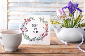 Card with words LOVE YOU MOM, cup of tea and flowers on table. Mother's day celebration