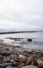 View of Salthill beach and Mutton island on a cloudy day, with rocks in the foreground and mountains in the background.