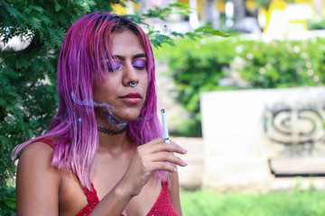 Relaxed young woman with makeup and pink hair, leaning against a tree smoking a cigarette. Concept of feminine beauty.